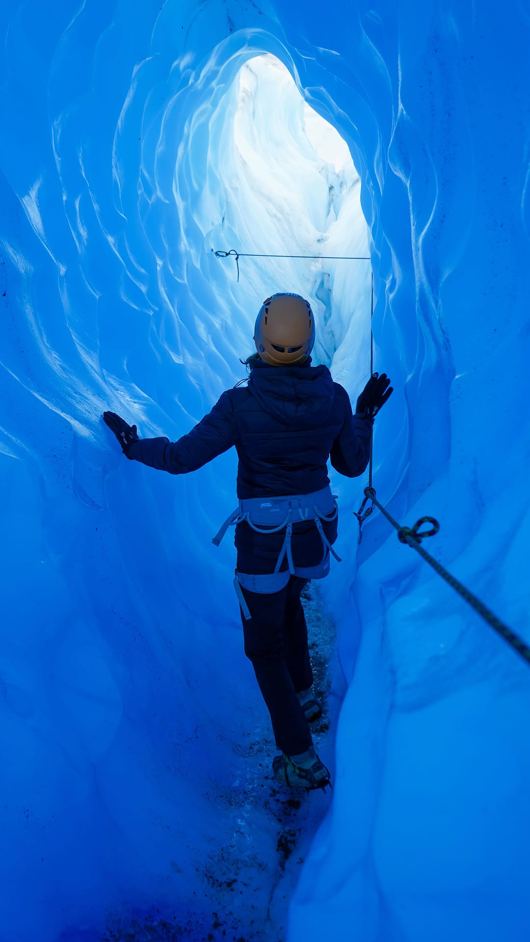 person walking inside narrow blue ice cave using safety rope and wearing climbing gear