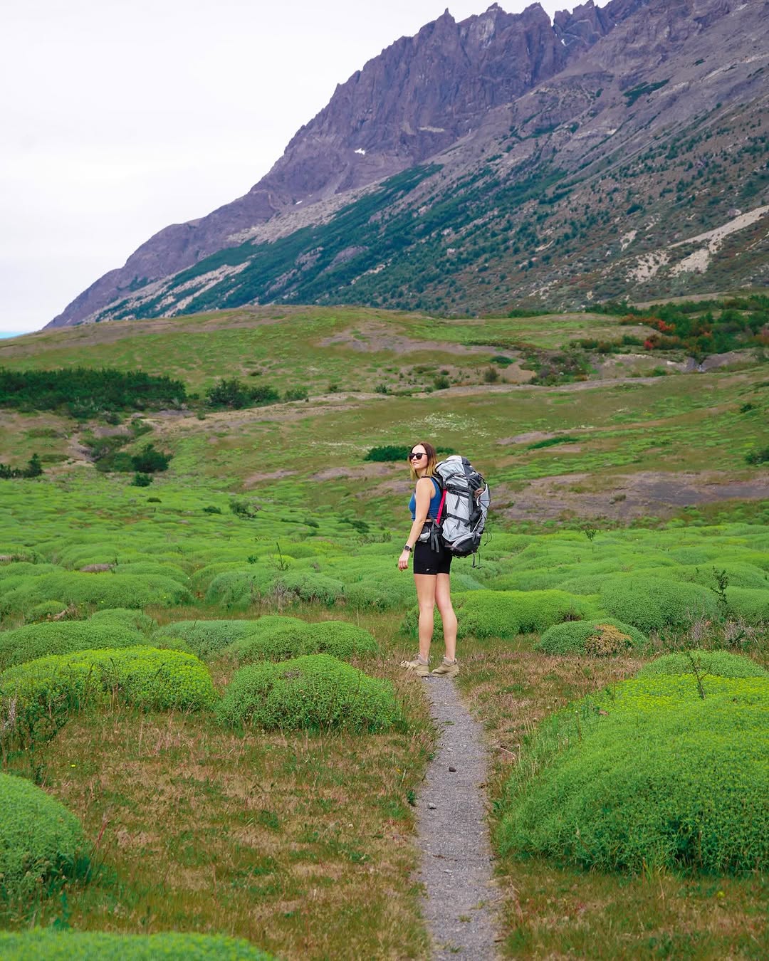 woman hiking on a narrow trail through green shrubbery with mountains in the background