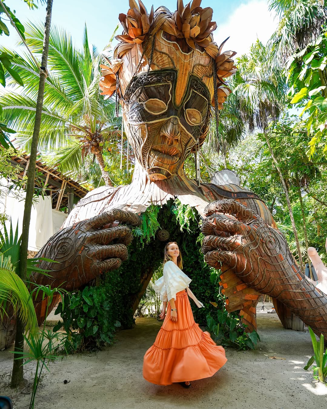 woman in a flowy orange skirt posing inside a large wooden sculpture of a person with plants