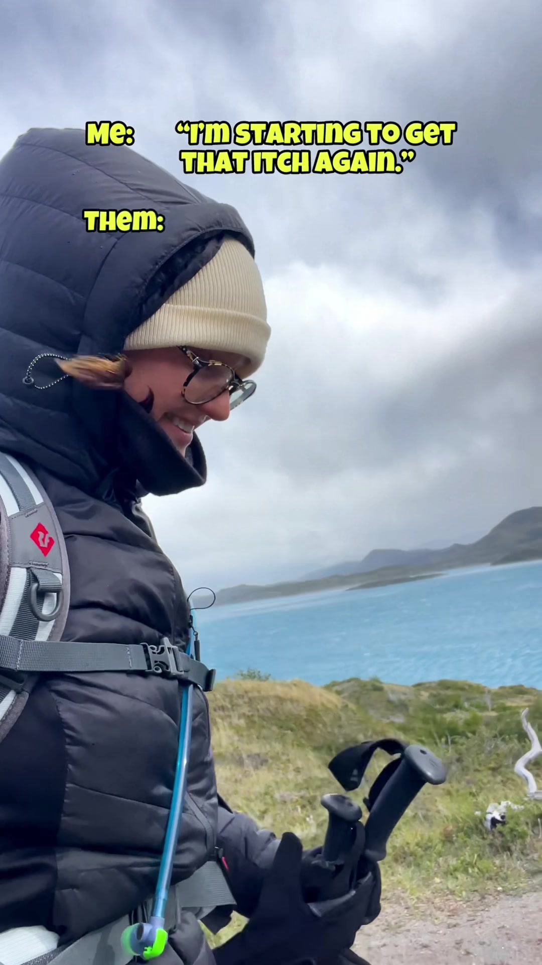person dressed for cold weather holding hiking poles by a blue lake under a cloudy sky
