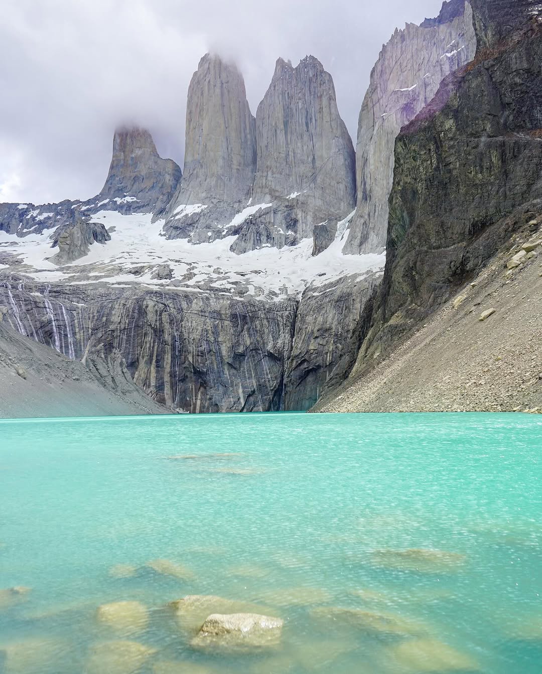 turquoise lake with rocky and snowy mountain cliffs under a cloudy sky