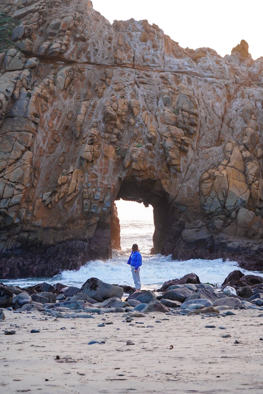 Person standing by a rock arch on a beach.