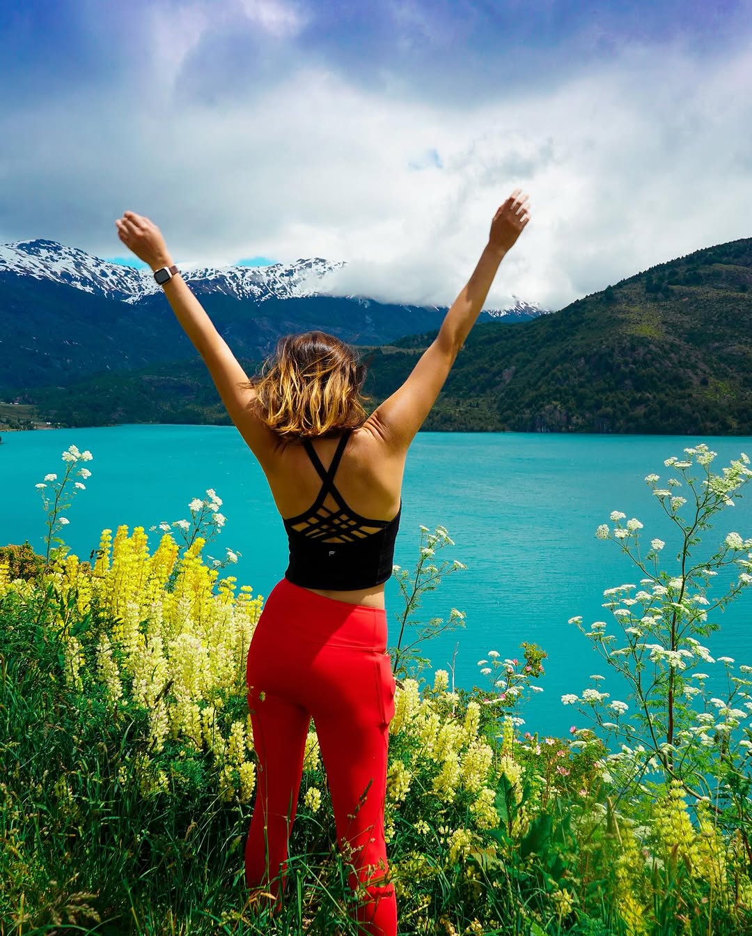 person with arms raised overlooking turquoise lake with mountains and yellow flowers