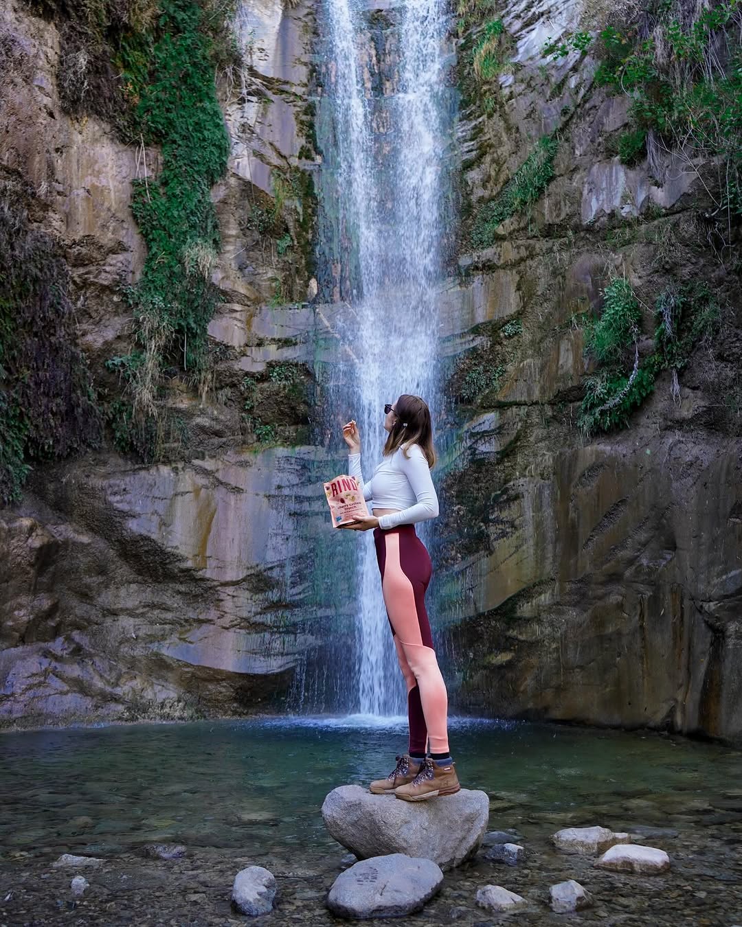 Polina standing on a rock in front of a waterfall, holding a bag of RIND snacks