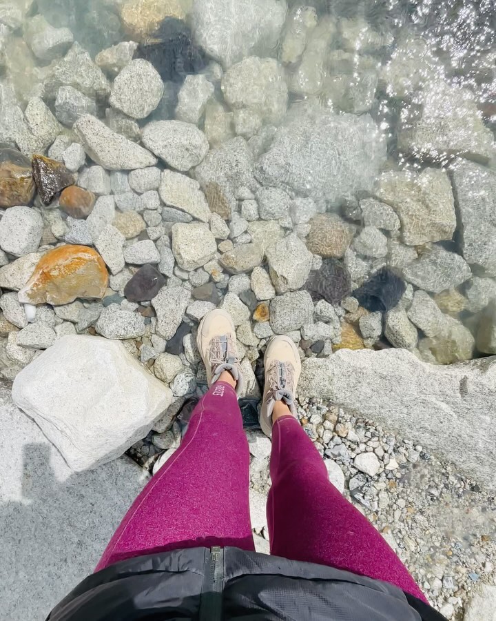 person wearing pink leggings and beige shoes standing on rocky shore with clear water