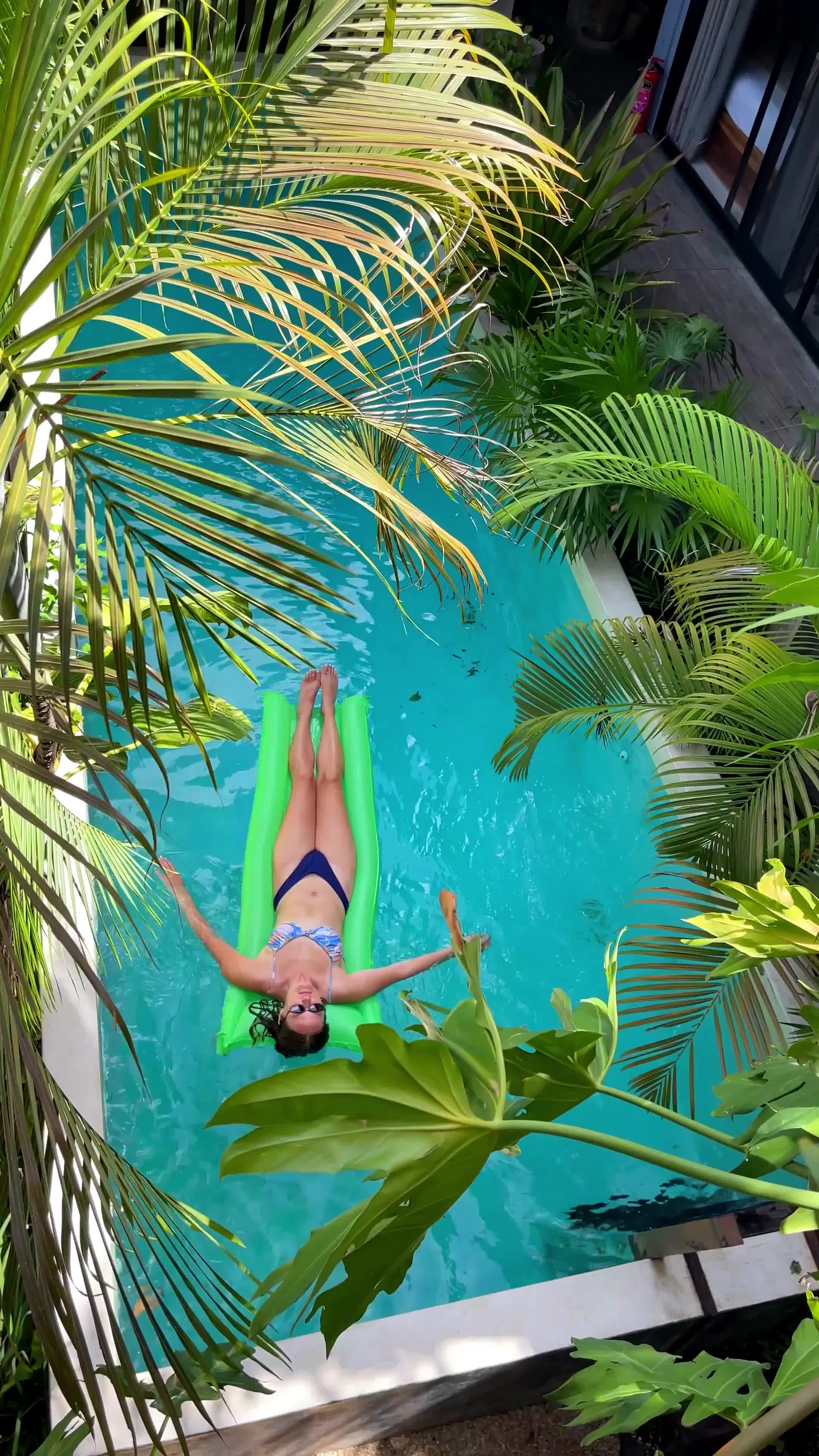 woman relaxing on a green float in a pool surrounded by tropical plants