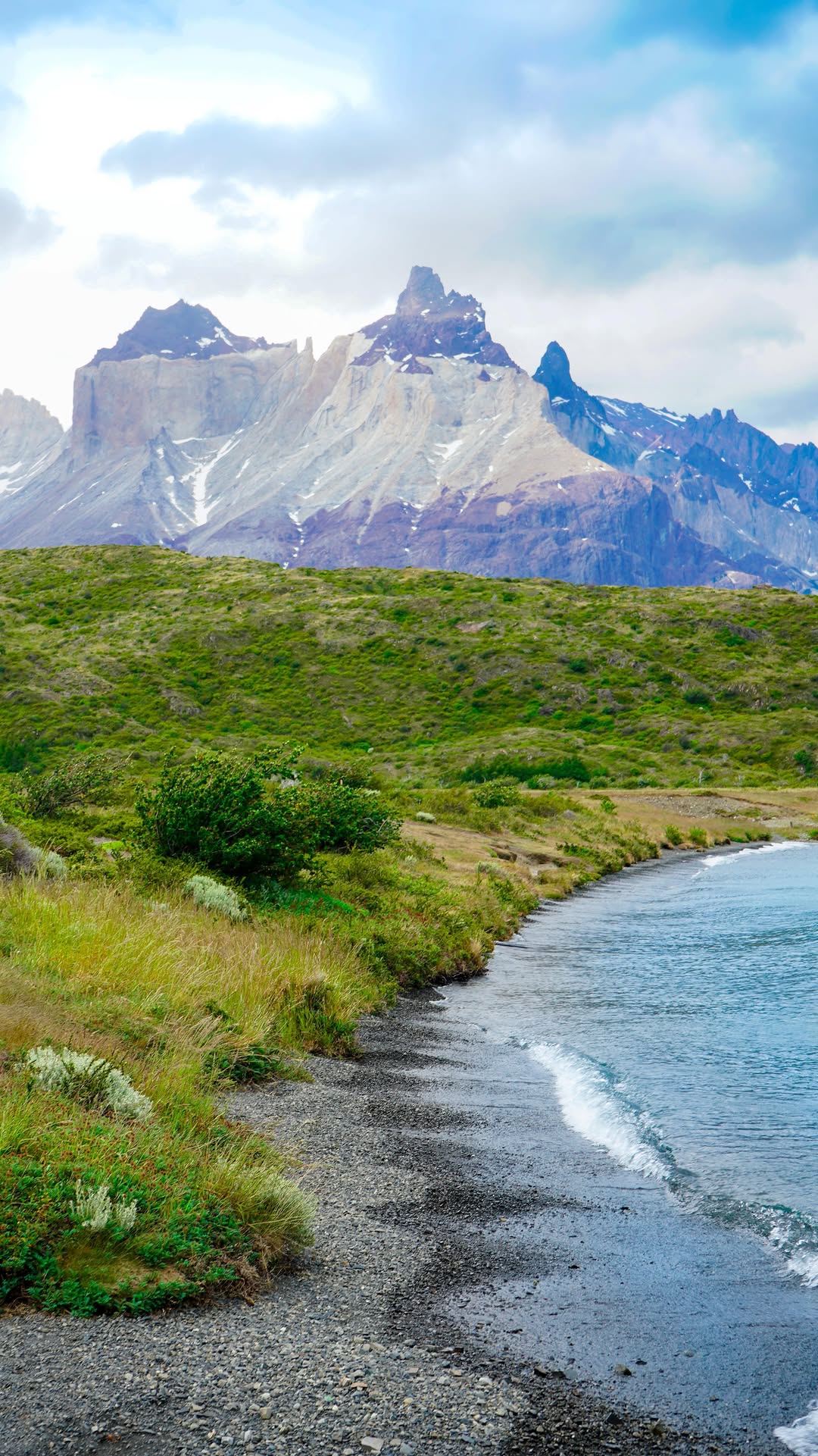 rocky shoreline with green grass and towering jagged mountains in the background under cloudy sky