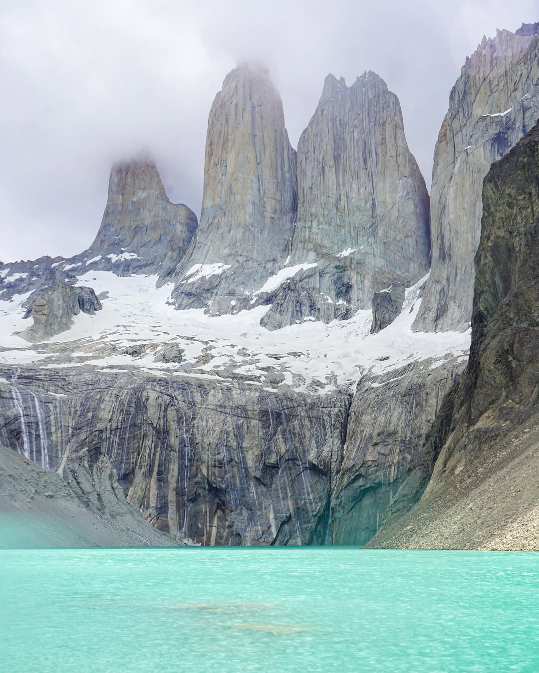 turquoise lake with towering rocky mountain peaks partially covered by snow and mist