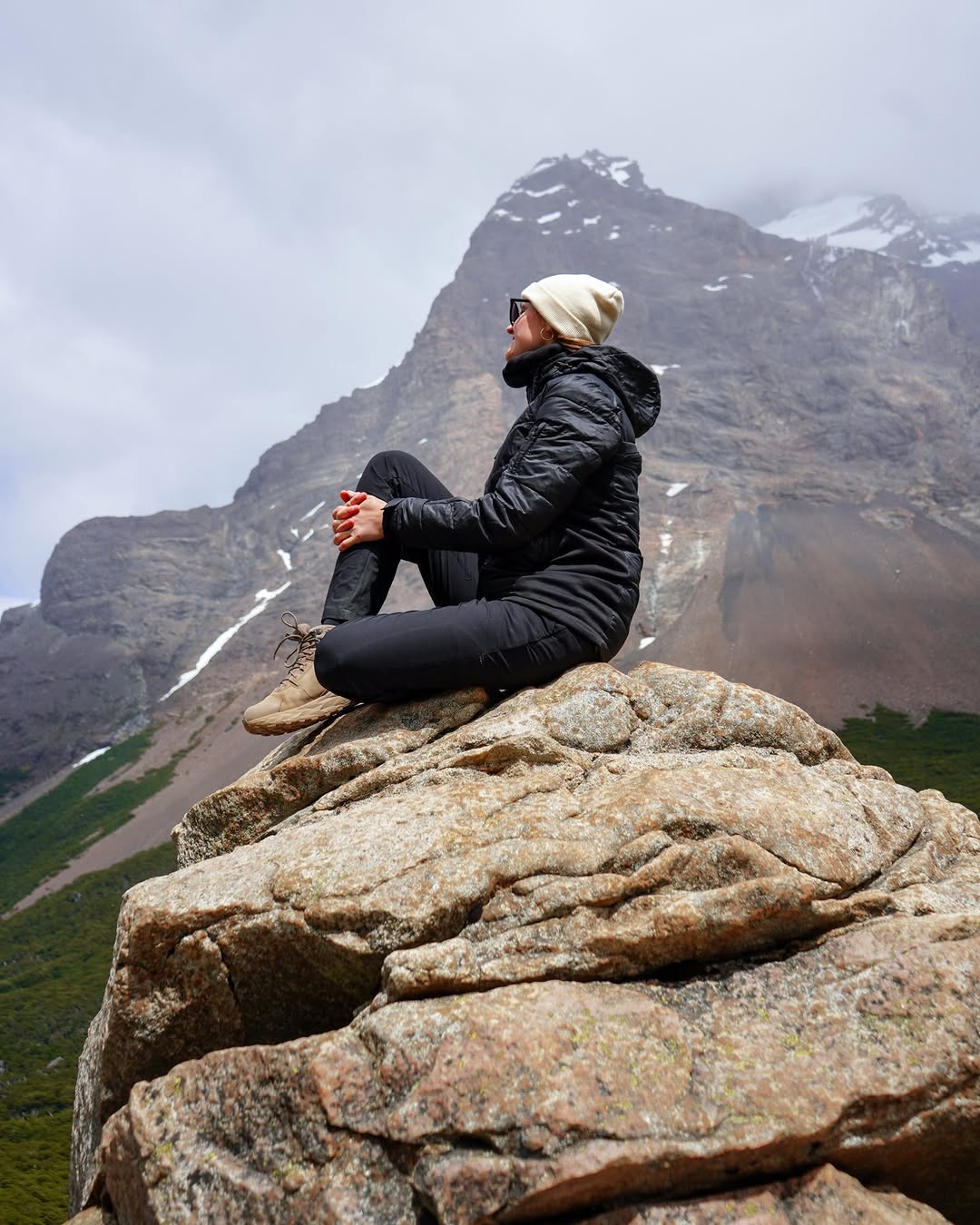 person sitting on rocky outcrop wearing winter clothes with mountain background