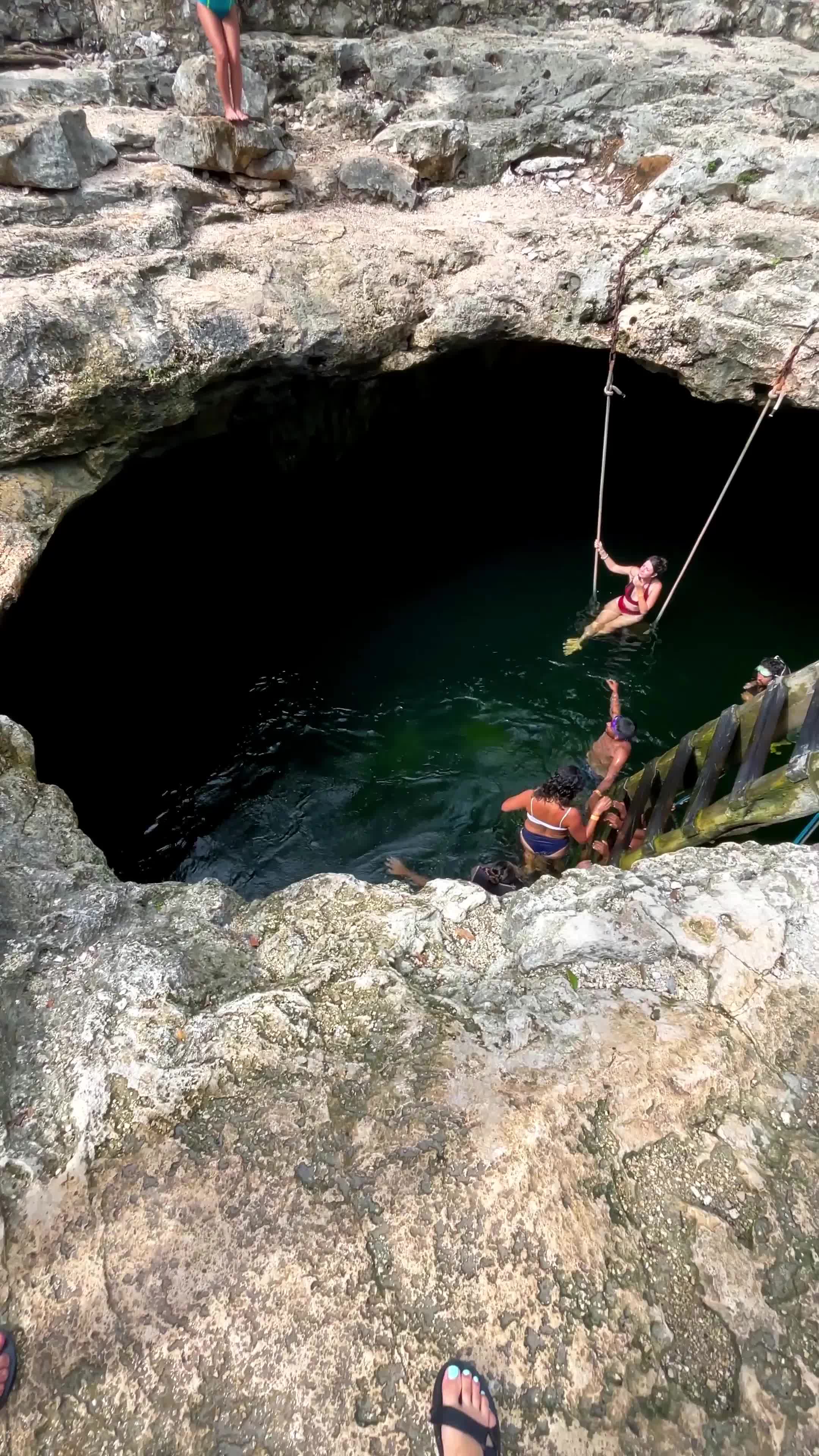 people swimming and swinging on ropes in a rocky natural pool or cave opening