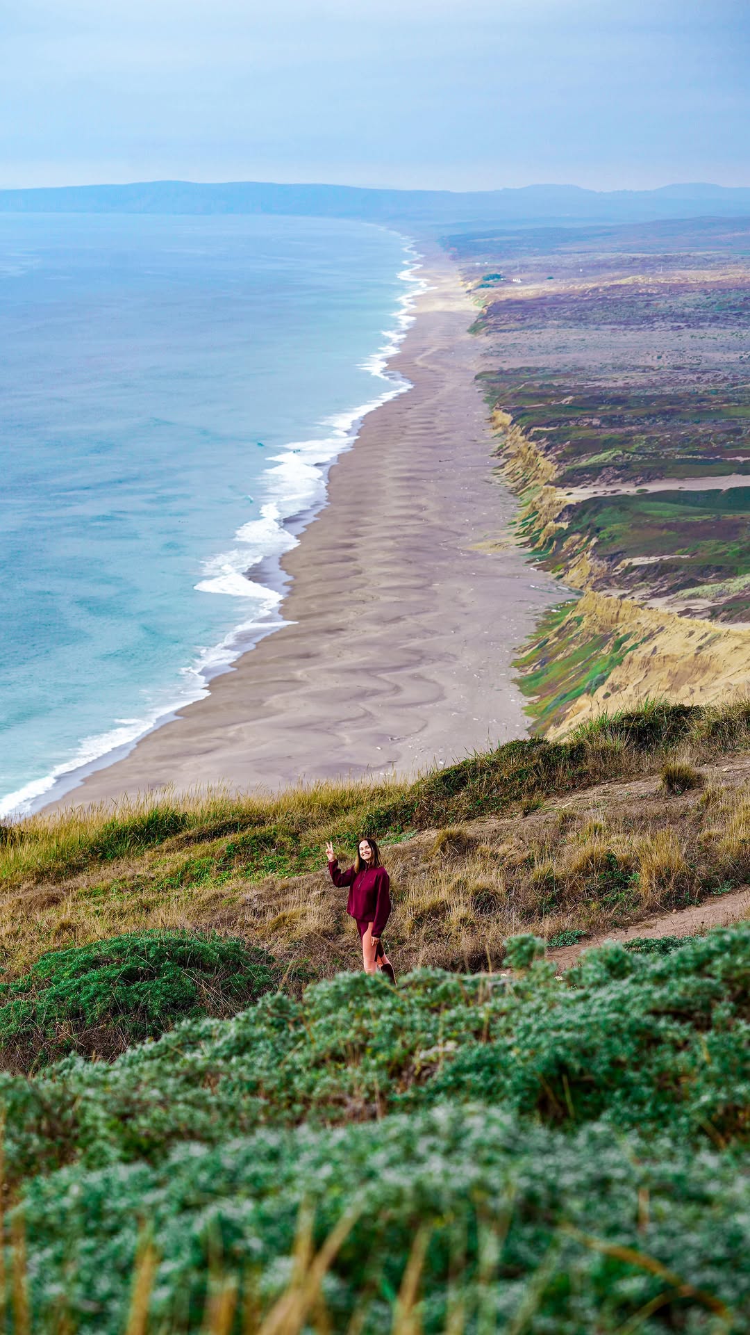 woman standing on a grassy hill overlooking a long sandy beach and ocean coastline