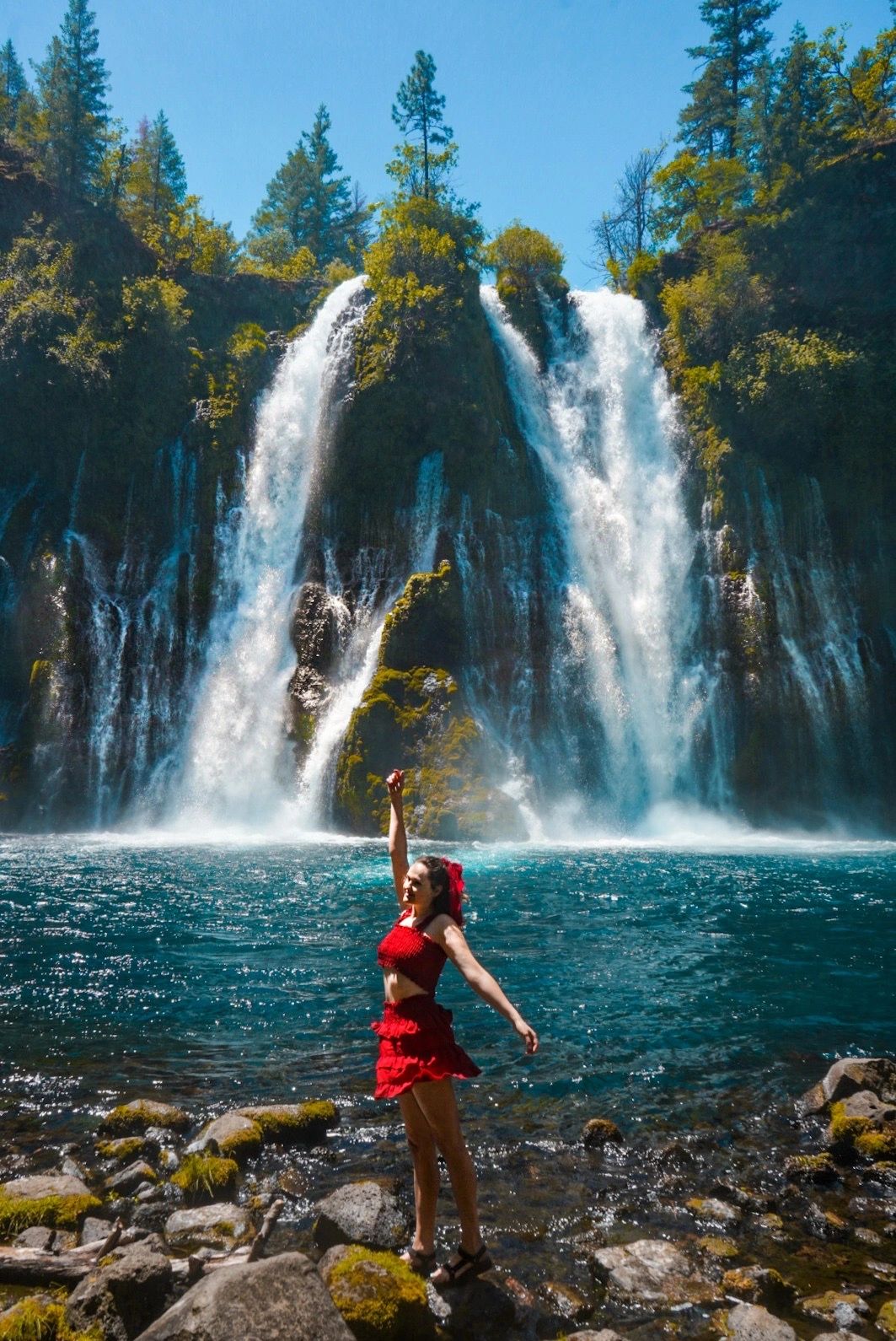 Mujer disfrutando frente a una cascada en un día soleado.