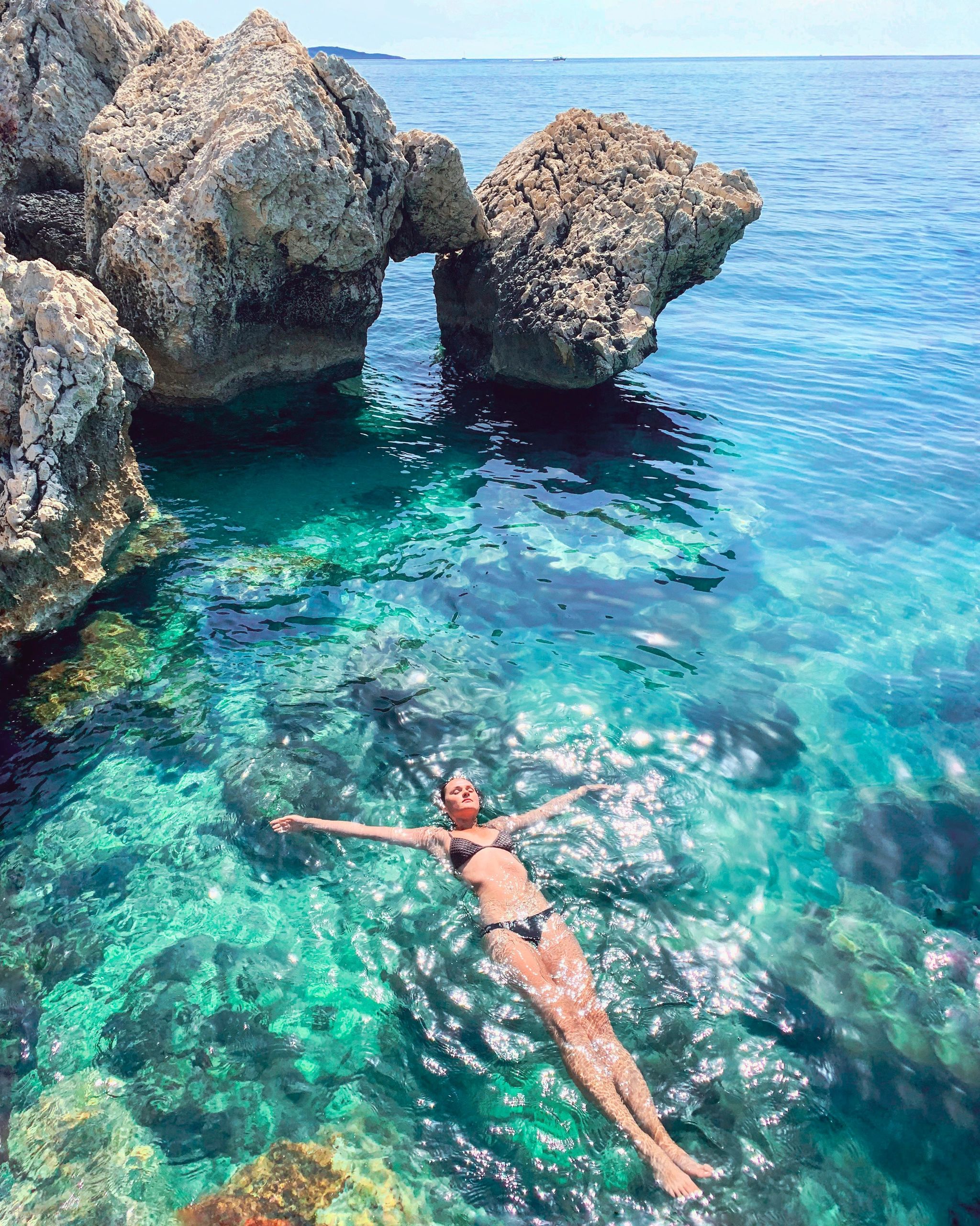 A girl floating in the green sea on summer day. Coast line is rocky and one rock looks like an arch.