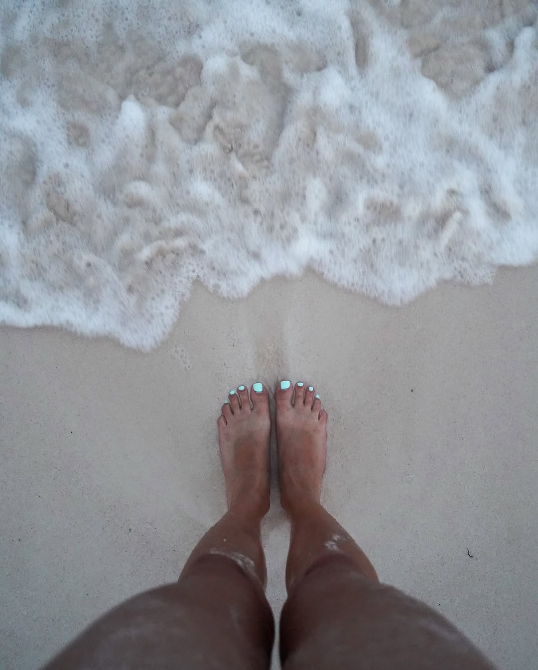 feet with turquoise nail polish standing on sandy beach near foamy ocean waves