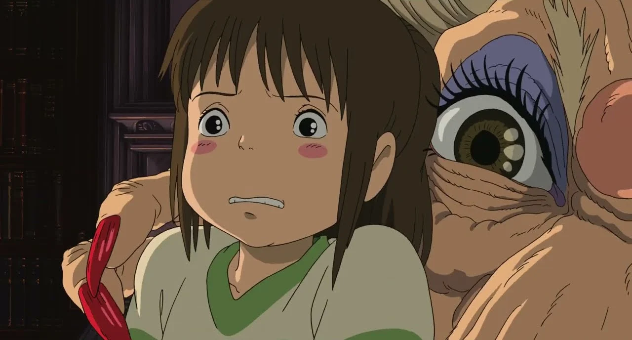 Chihiro looks frightened as Yubaba leans in close beside her in a scene from Spirited Away.