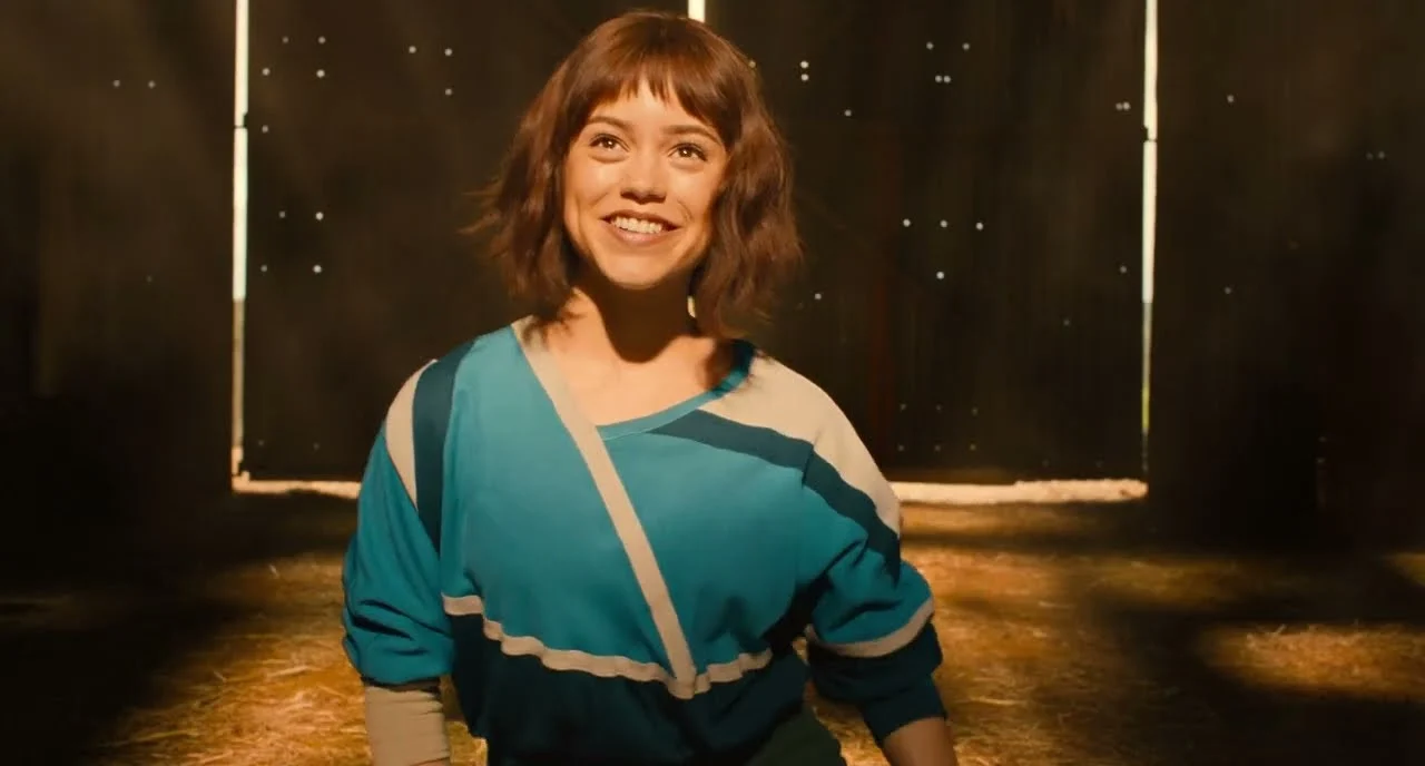 A smiling young woman in a teal jacket stands in a barn lit by low sun in a scene from Klara and the Sun.