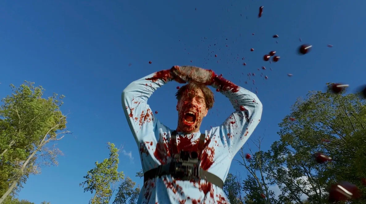 Still from REBRAND: a man in a blood-spattered shirt screams with his hands on his head under a blue sky