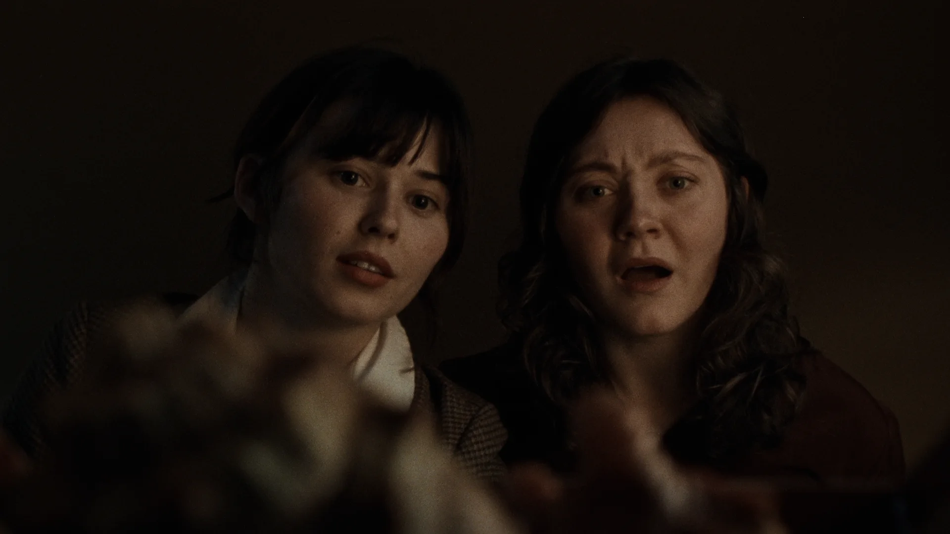 Still from STEAK DINNER: two women lean in close and stare down in alarm in a dim room