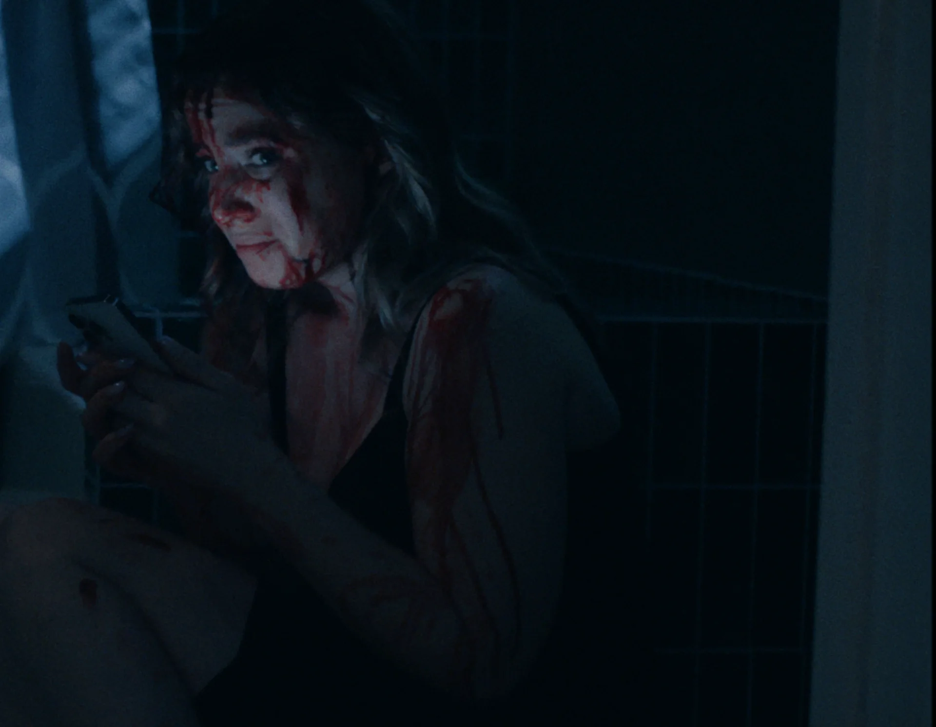 Still from STAR-CROSSED: a woman with blood on her face sits on a bathroom floor at night looking at her phone