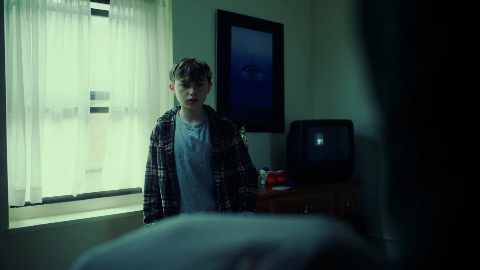 Still from ZIPPY'S HOUSE: a teenage boy in a plaid shirt stands in a dim bedroom beside an old television