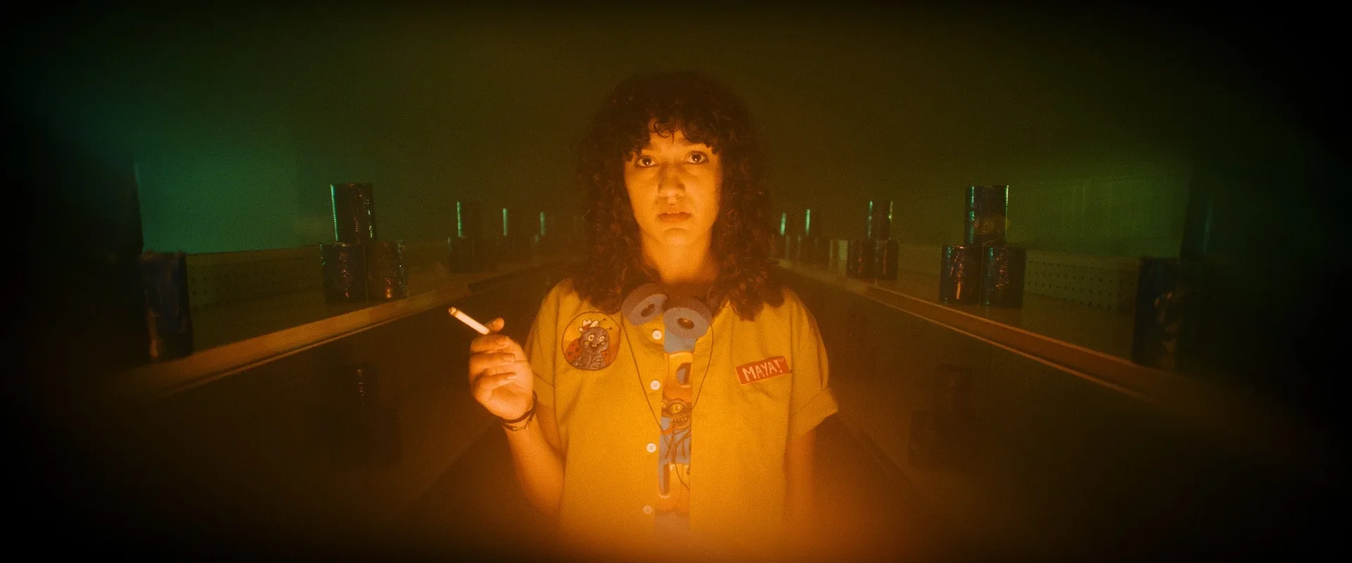 Still from DEAD ENDERS: a clerk with curly hair holds a cigarette in a room lit orange and green