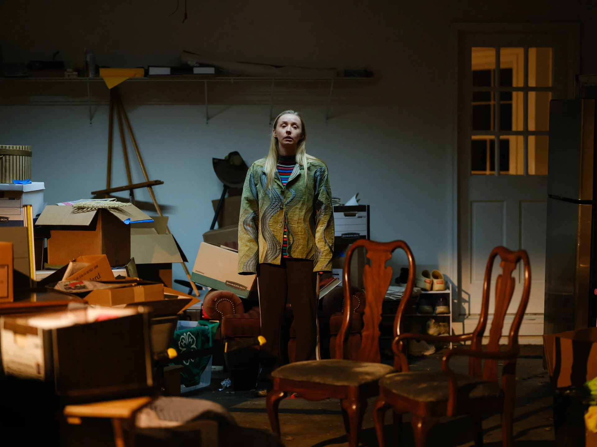 Still from LOVE IN ALL THE WRONG PLACES: a woman stands in a cluttered room among dining chairs and boxes