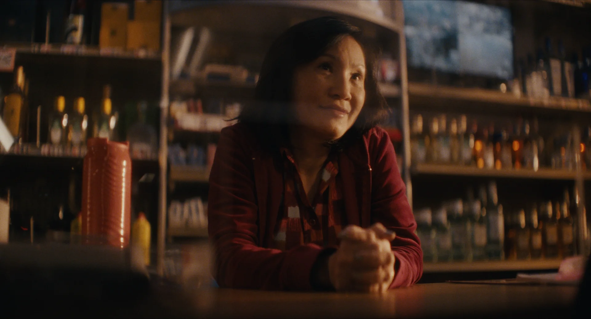 Still from LONG’S LONG LOST & MINI MART: an older woman smiles with her hands clasped at the counter of a convenience store