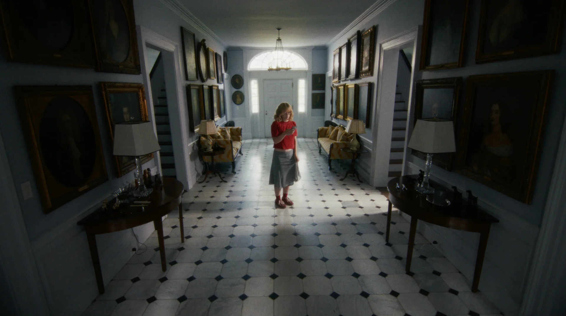 Still from OH SOLO VIOLET: a woman in a red top stands alone in a long hallway with a checkered floor and framed paintings