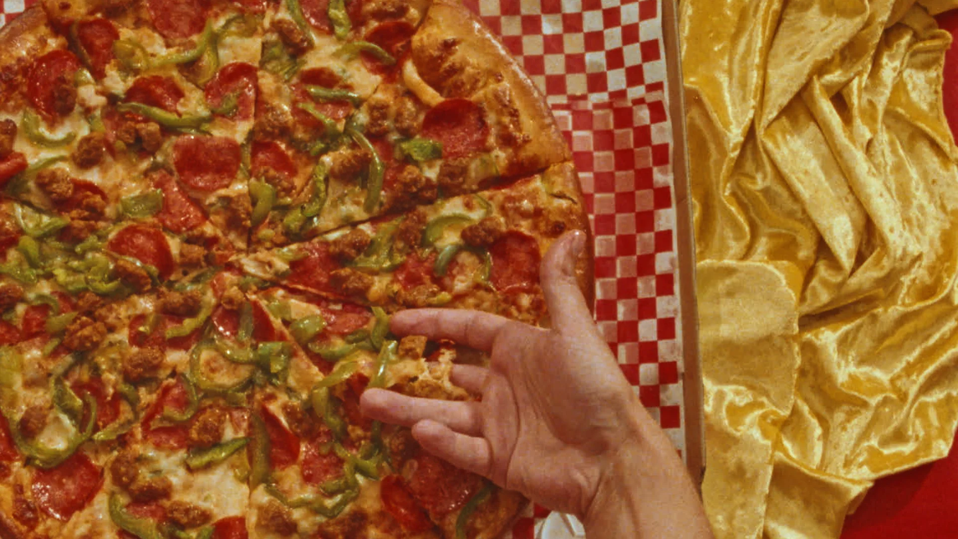 Still from MAKE ME A PIZZA: a hand reaches for a pepperoni and green pepper pizza beside red checkered paper and gold fabric