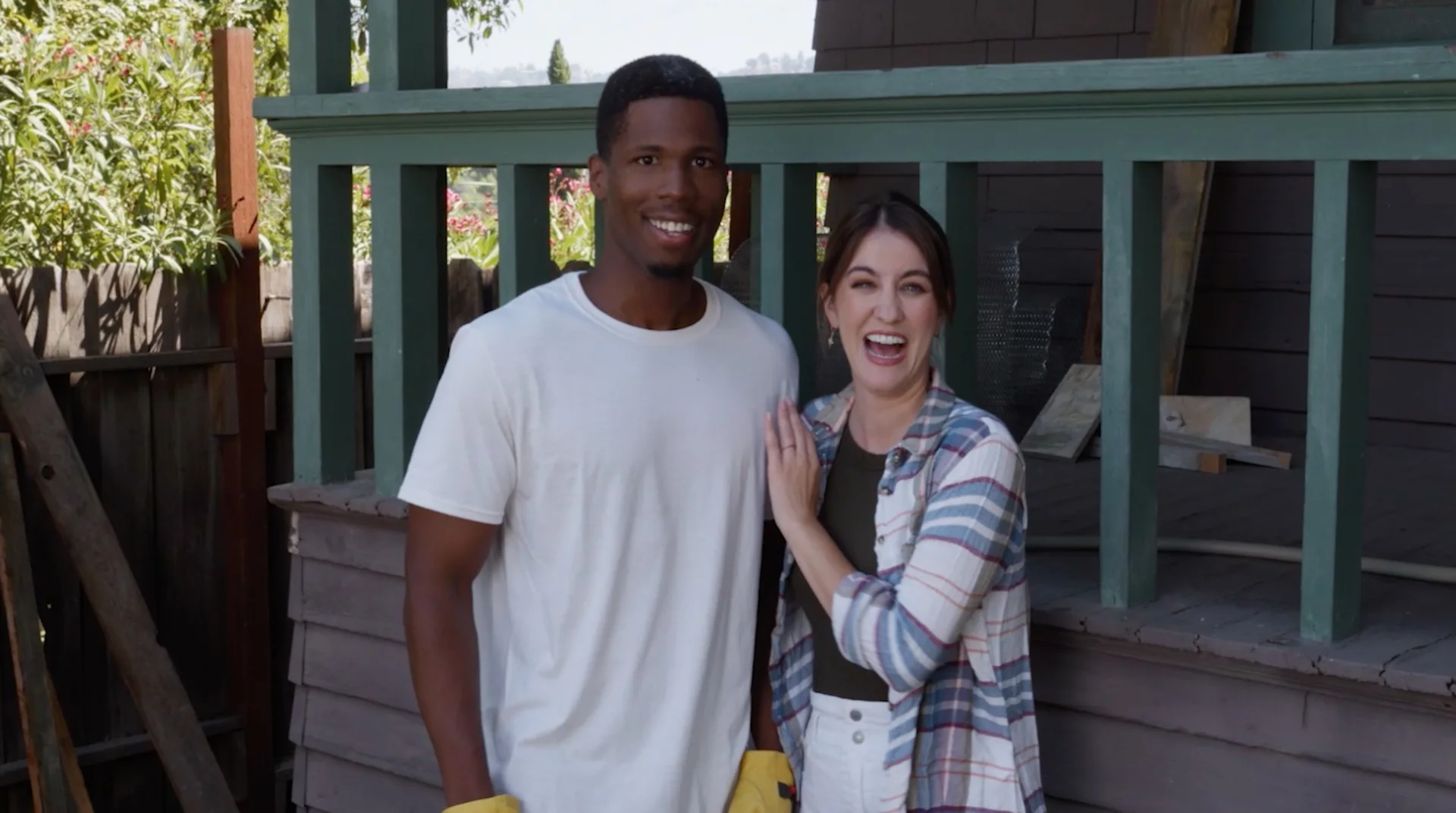 Still from THE LAST CHEAP HOUSE: a smiling couple in work gloves stand in front of a house under renovation