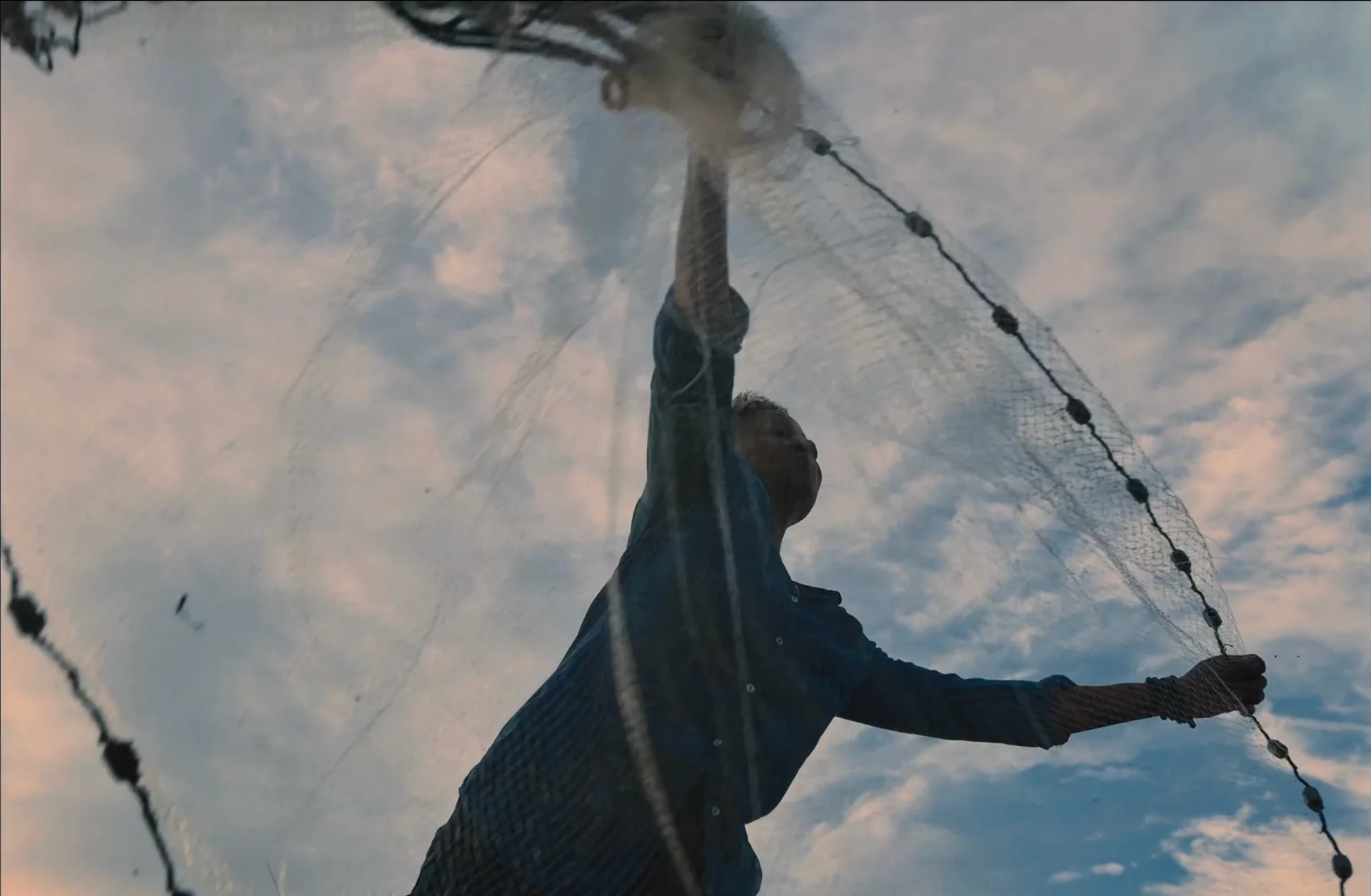 Still from HAINT: a person casts a fishing net into the air against a cloudy sky