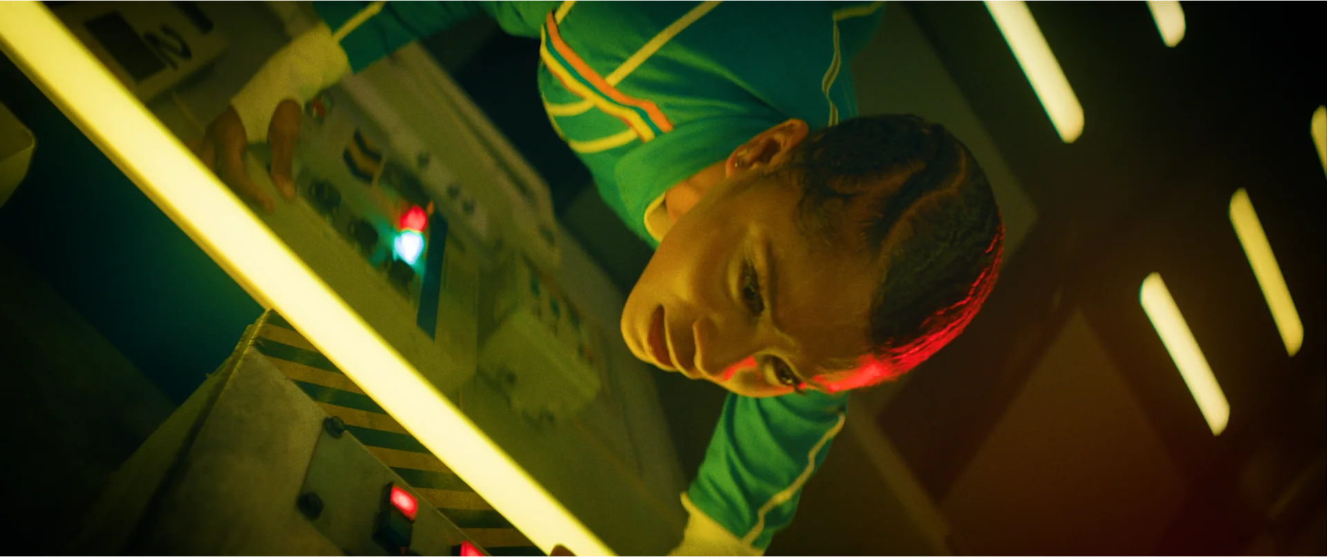 Still from TETHER: a woman in a green jacket, seen at an angle in a corridor lit with yellow lights