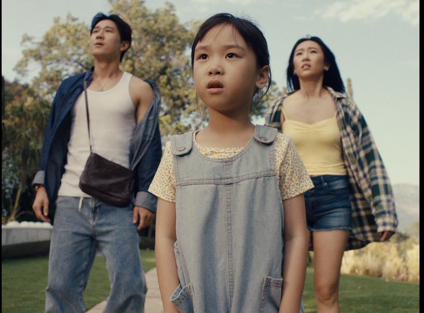 Still from RESET: a man, a young girl in overalls and a young woman stand outdoors looking at something out of frame