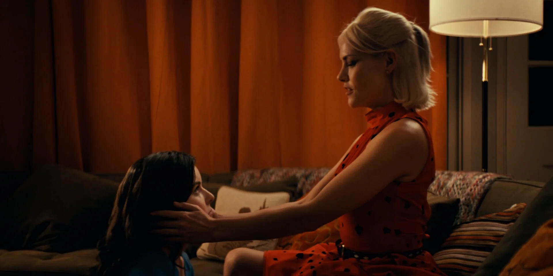 Still from OPEN WIDE: a blonde woman in a red dress holds the face of a dark-haired woman on a sofa in an orange-lit room