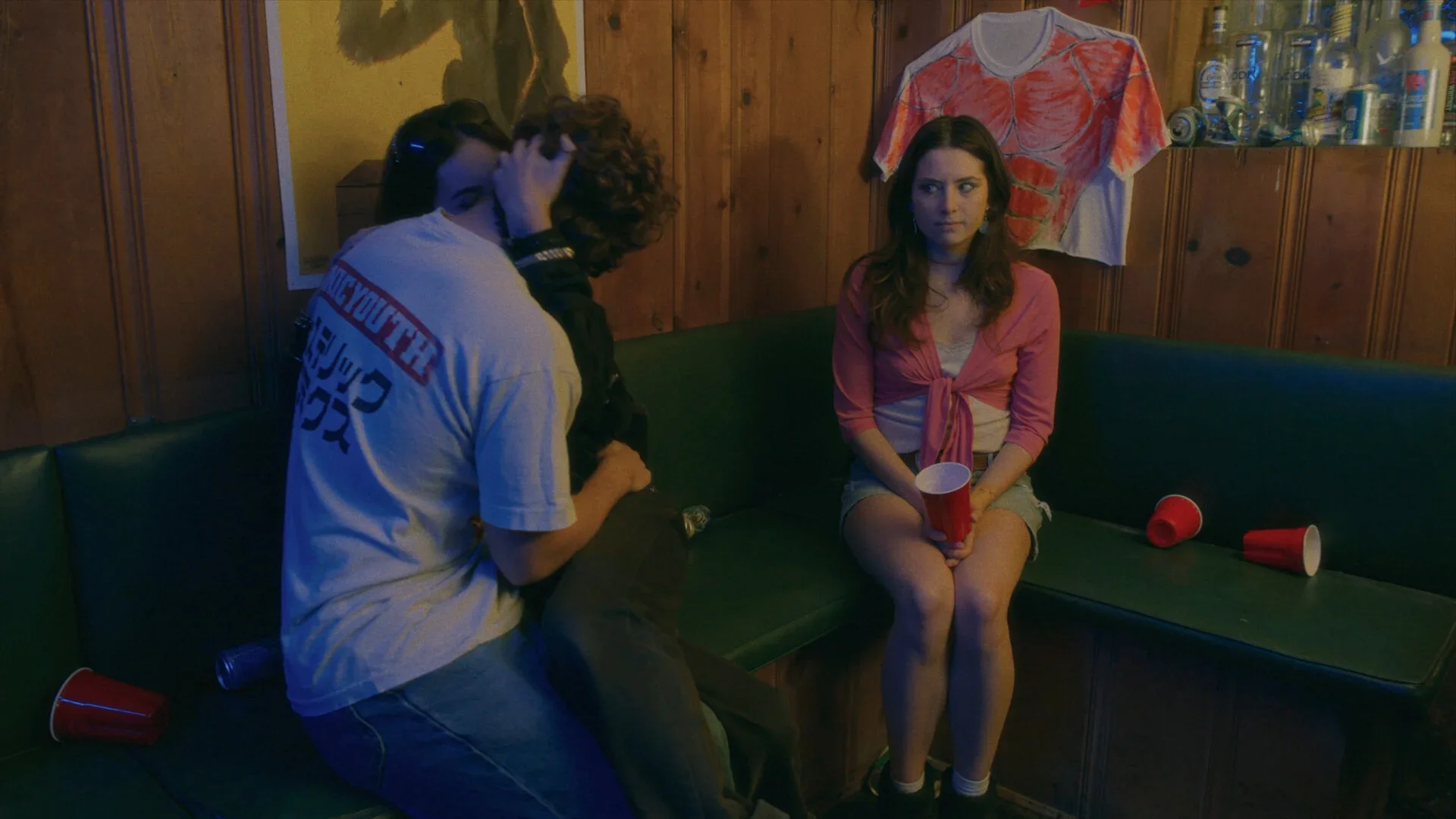 Still from CHAMELEON: a teenage girl sits on a booth seat holding a red cup beside a couple kissing