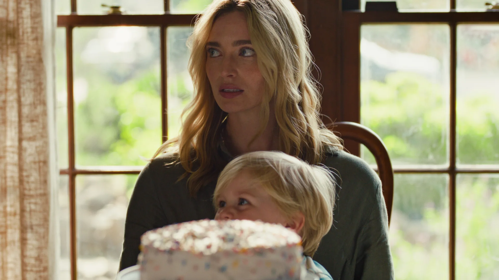 Still from BENNY'S SECOND BIRTHDAY: a blonde woman holds a toddler on her lap behind a birthday cake, looking off to the side