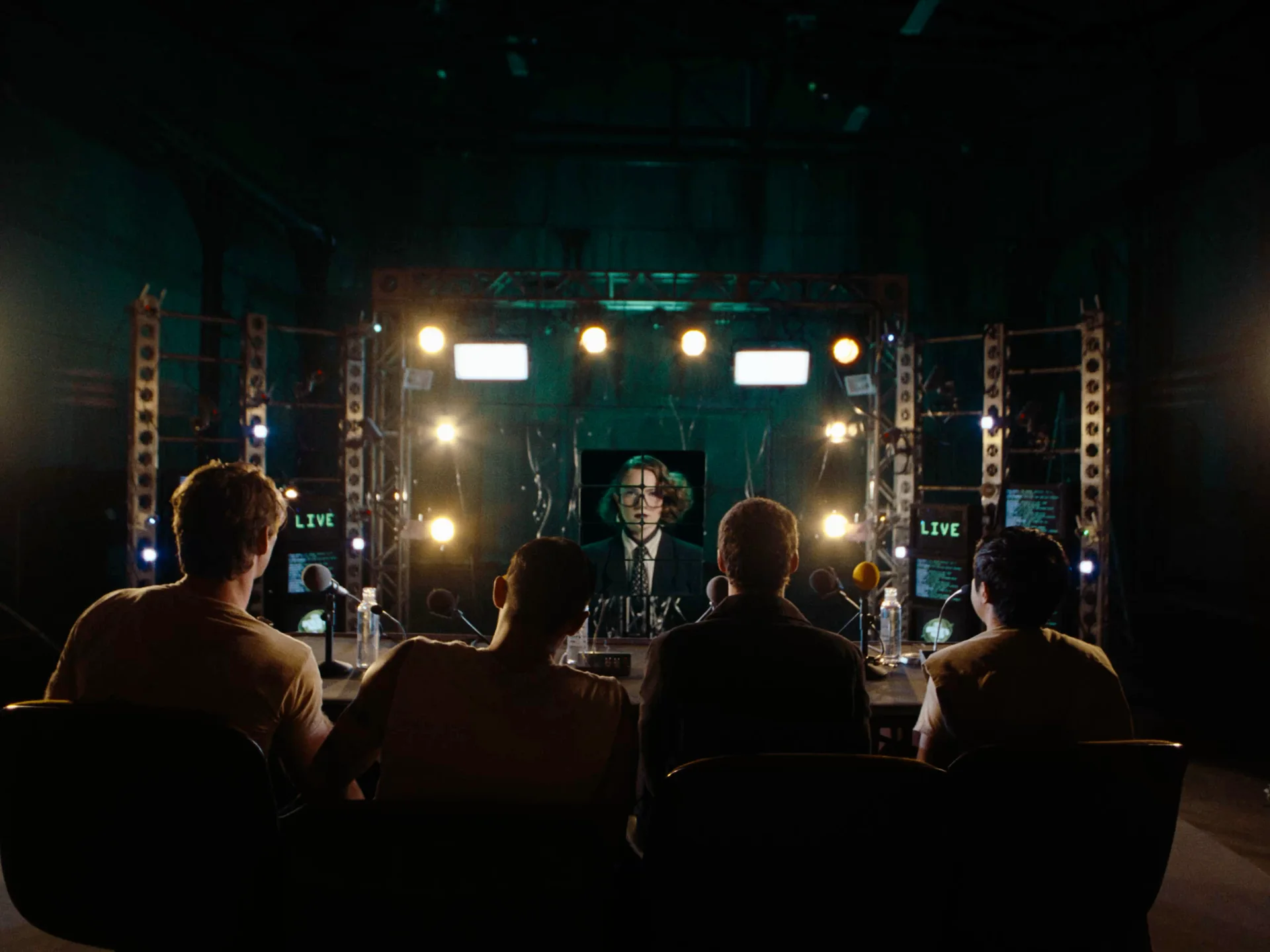 Still from FOUR ITCHY BOYS: four men seen from behind sit at a table facing stage lights and a screen