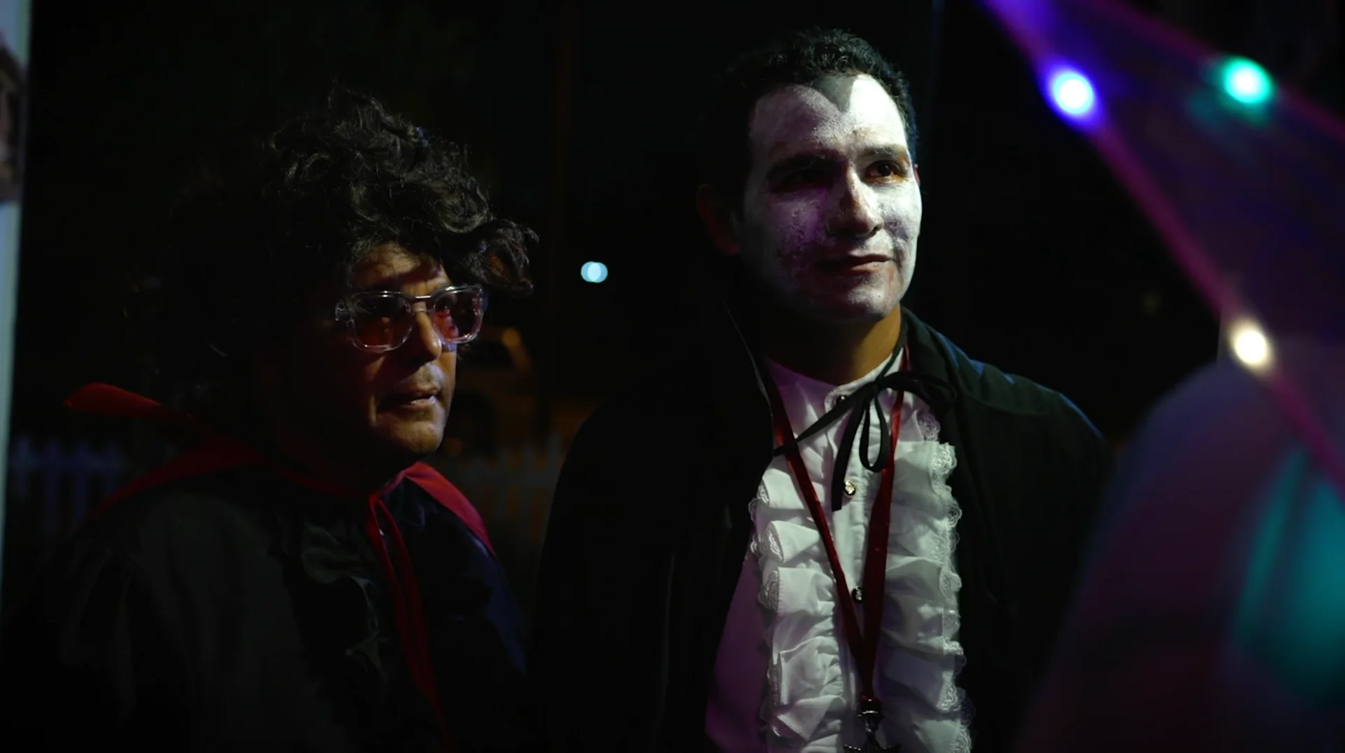Still from VAMPIRE FOR HIRE: a man in pale vampire makeup and a ruffled shirt stands beside a man in glasses and a curly wig