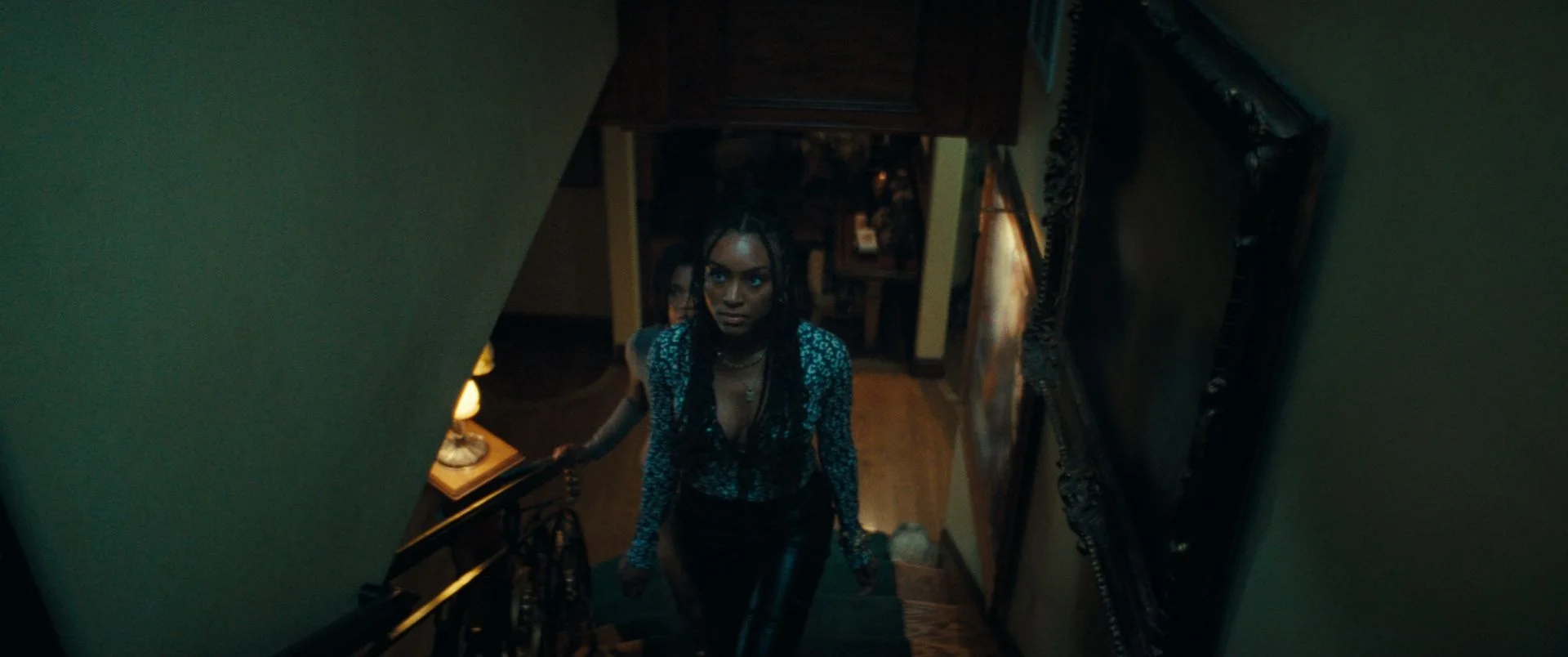 Still from THE BRAID: a woman looks up from a staircase in a dim, green-lit house