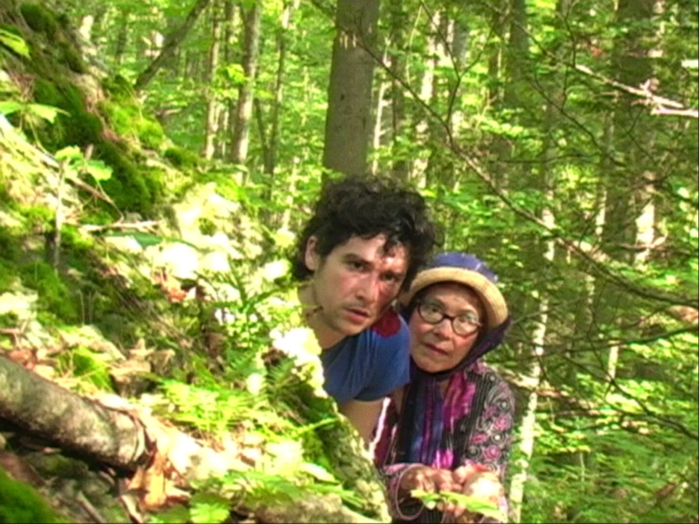 Still from HAROLD EATS MAUDE: a young man and an older woman in a hat crouch together in a sunlit forest