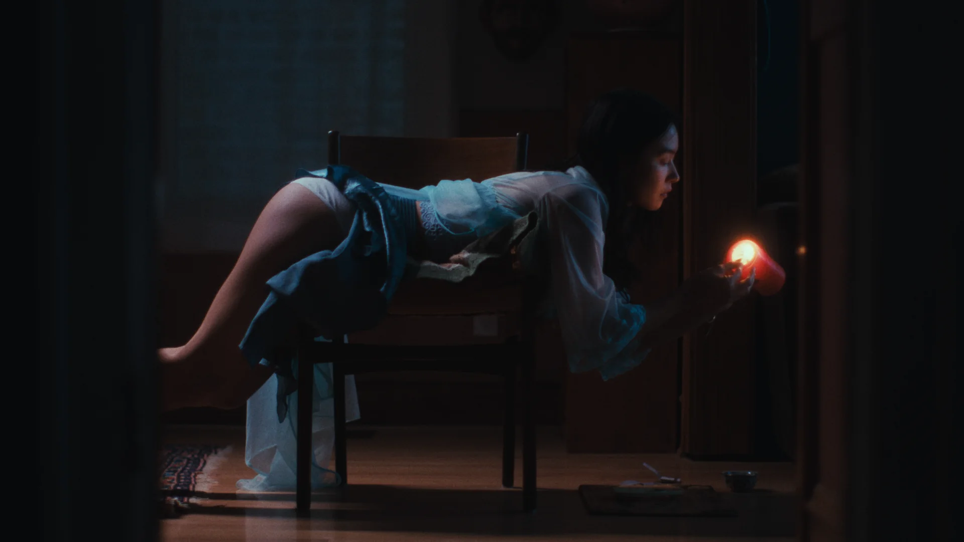 Still from SPANKED BY A GHOST: a woman lies face down across a wooden chair in a dark room, holding a lit red candle