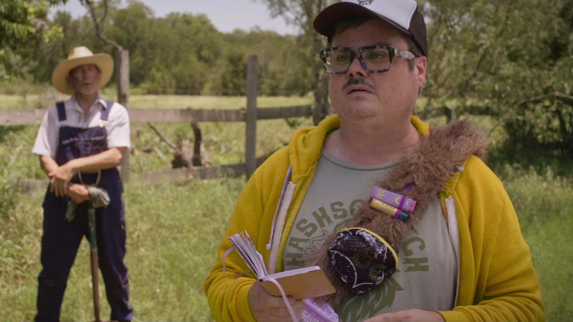 Still from IT'S HARD NOT TO BE ROMANTIC ABOUT TIME TRAVEL: a man in a yellow hoodie, glasses and a trucker cap holds a book in a field, with a farmer in overalls behind him