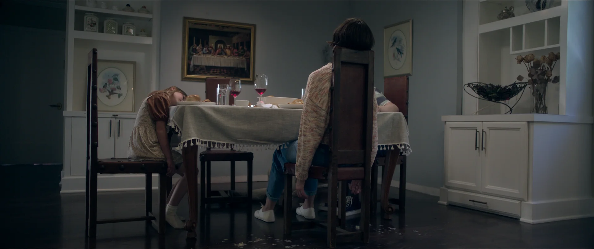 Still from THE SEQUENCE: two people sit at a dining table, one slumped face down, under a painting of the Last Supper