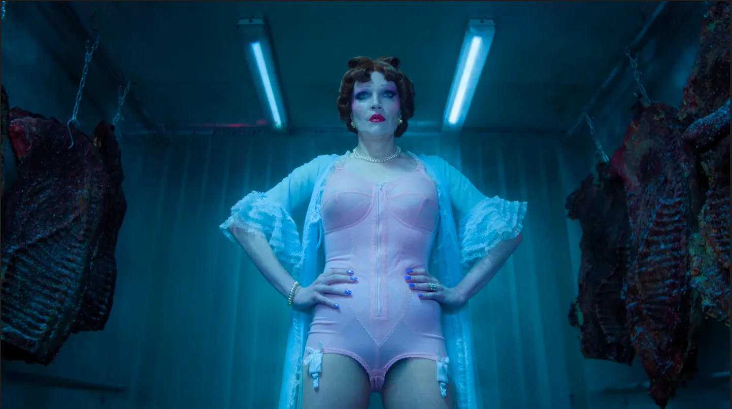 Still from CHARLIE IS NOT A BOY: a figure in pink lingerie, a pale blue robe and doll-like makeup stands with hands on hips in a blue-lit meat locker