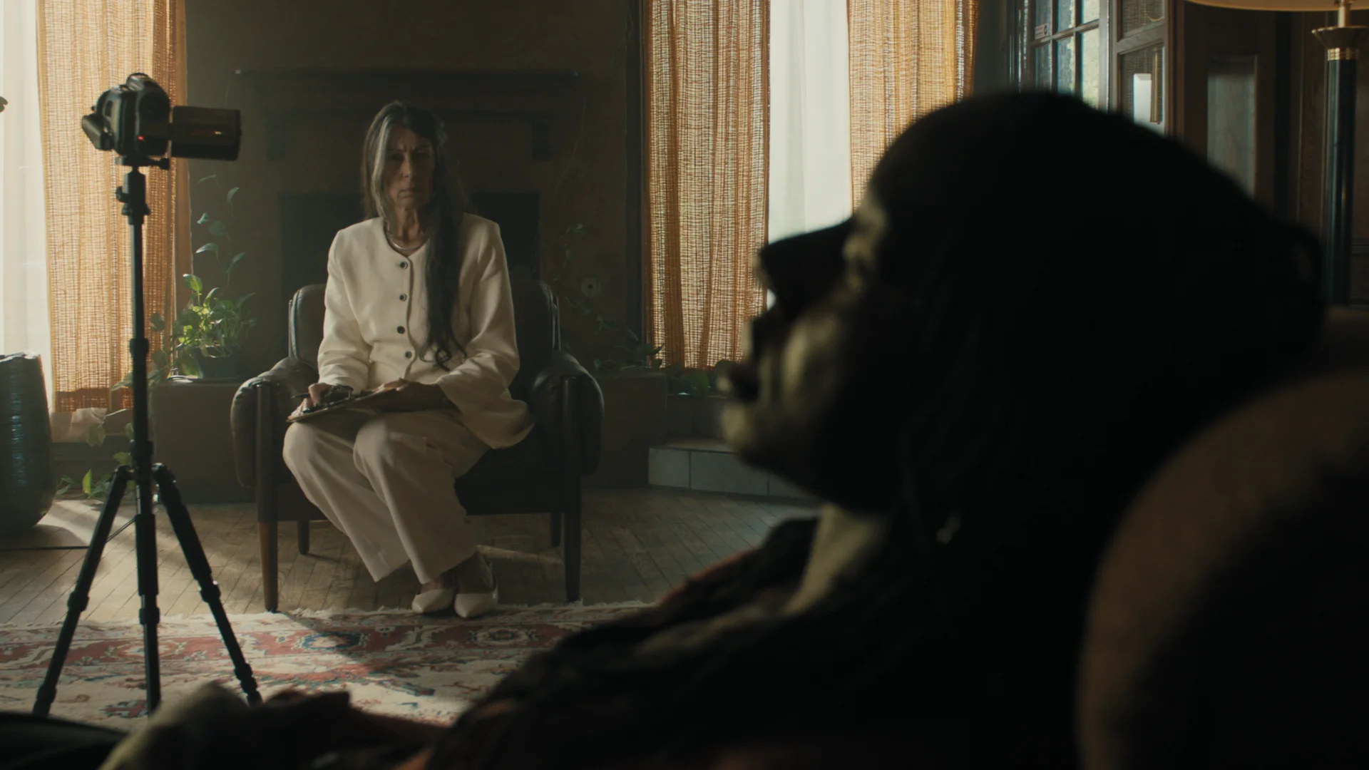 Still from TRANSFERENCE: a woman in white sits in an armchair beside a camcorder on a tripod, facing a person in the foreground